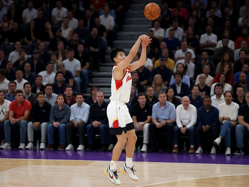 NBA篮球场上球员跳投瞬间，篮球在空中划出弧线，背景是模糊的观众席